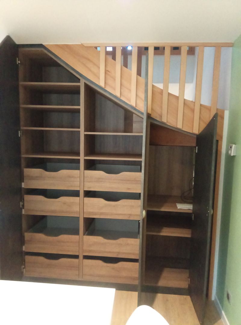 Le même ensemble ouvert : étagères, nombreux tiroirs à poignée découpée et penderie en décor chêne derrière des portes sombres, sous la volée d'escalier