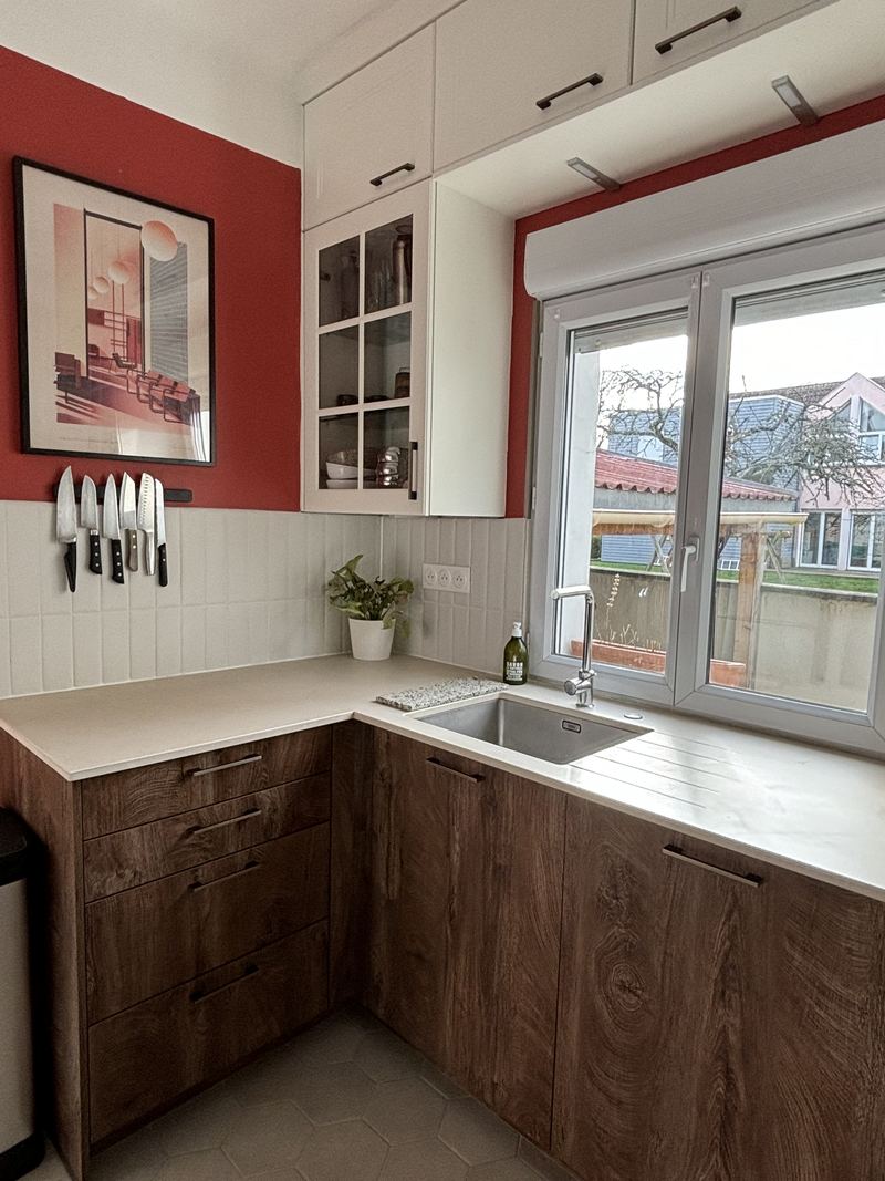 Angle évier de la même cuisine : meubles bas bois foncé, meubles hauts crème avec vitrine, barre aimantée à couteaux, crédence blanche, évier inox sous fenêtre à volet roulant