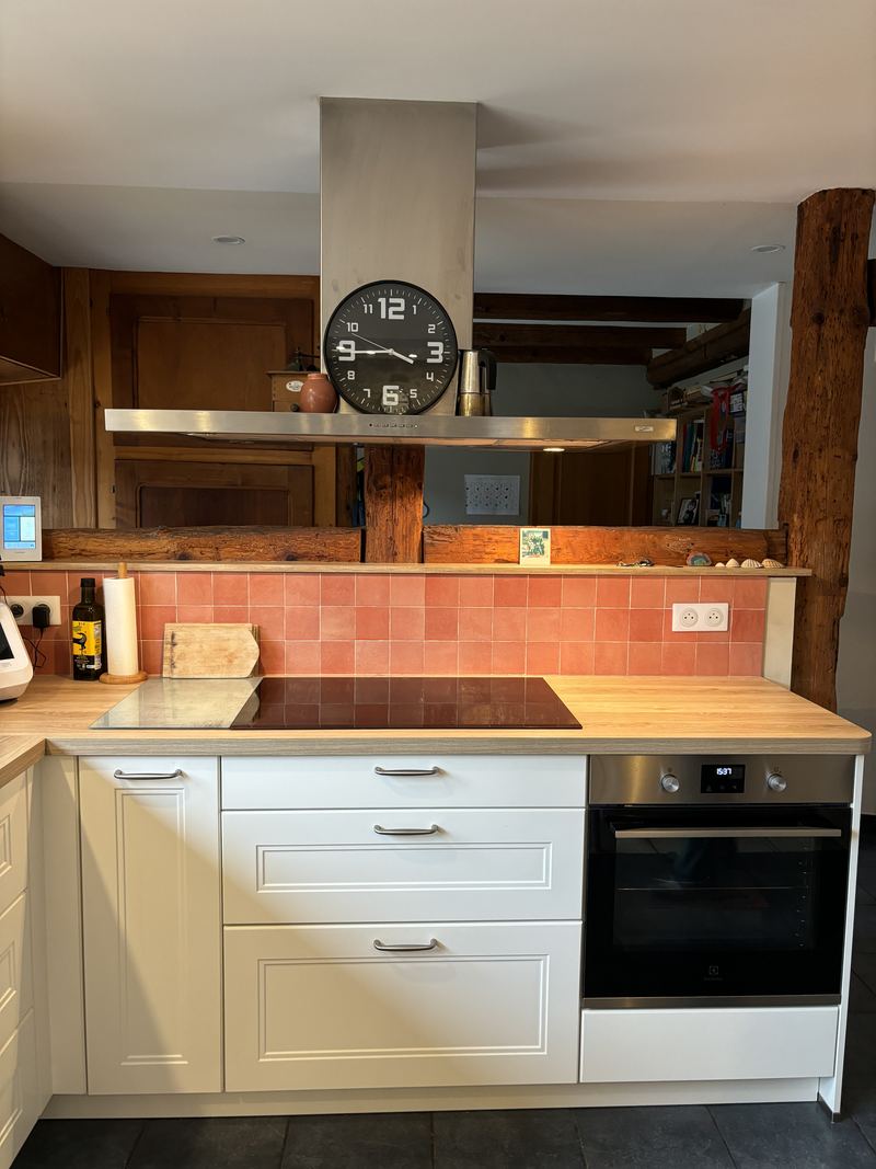 Plaque induction sous hotte îlot inox (horloge posée dessus), meubles crème à moulures et poignées métal, four inox encastré, plan décor bois, crédence en carreaux terracotta