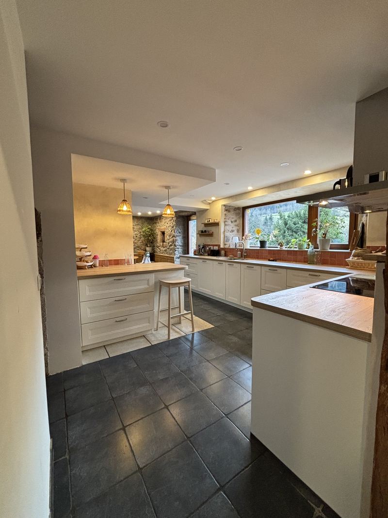 Vue d'ensemble de la cuisine crème à moulures avec retour, plan décor bois, crédence terracotta, faux plafond à spots, mur en pierre apparente et grande fenêtre sur la verdure