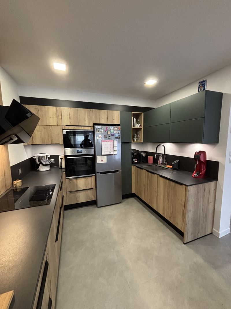 Cuisine en U : façades décor chêne noueux à poignées noires, meubles hauts vert sapin mat, plan de travail noir, hotte inclinée noire, fours encastrés, réfrigérateur inox pose libre