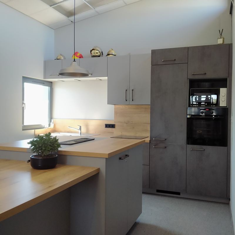Cuisine grise : meubles bas et hauts gris clair, colonne décor béton (four, micro-ondes), plan de travail et crédence décor chêne clair, évier sous fenêtre