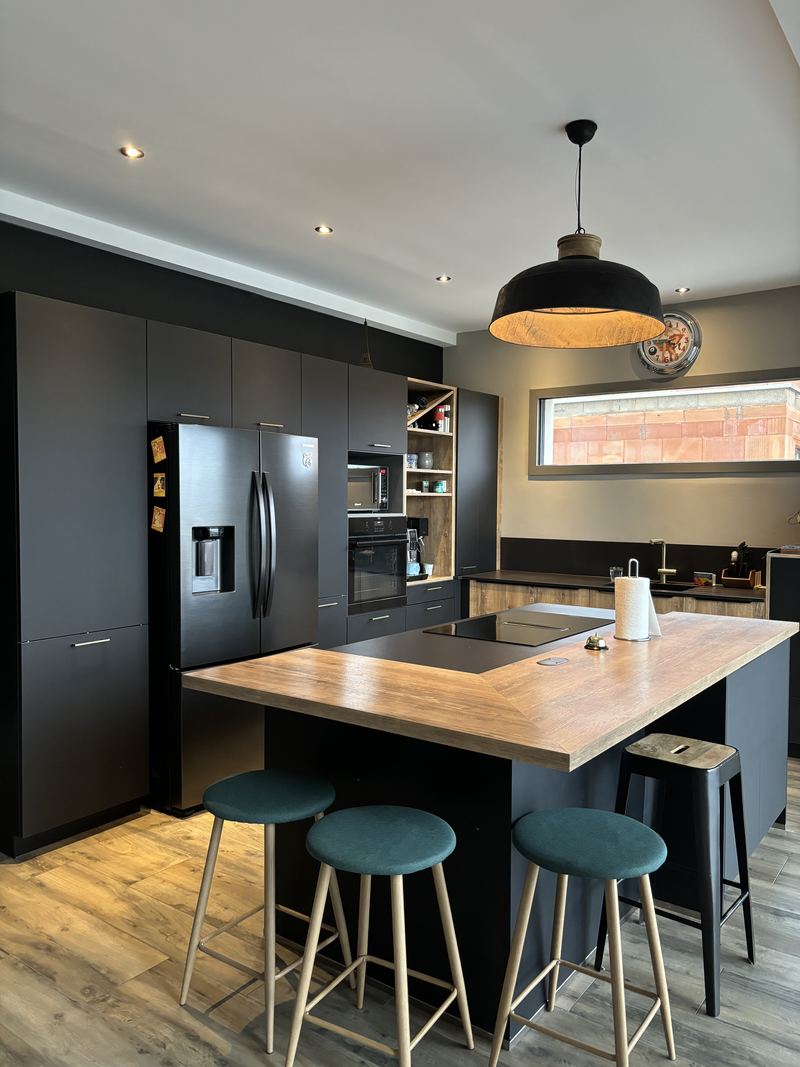 Vue d'ensemble : îlot noir à plan de travail décor bois débordant avec trois tabourets à assise bleu canard, colonnes noires avec réfrigérateur américain, grande suspension noire, fenêtre en bandeau