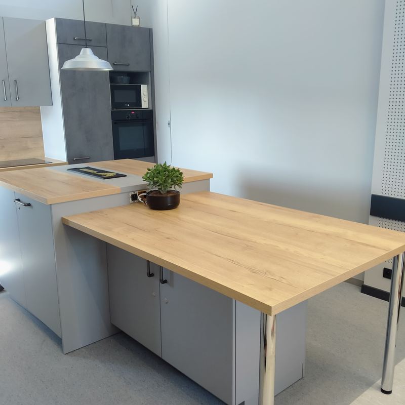 Table en décor chêne clair sur pieds chromés prolongeant un îlot gris clair, colonne décor béton avec four et micro-ondes, suspension blanche