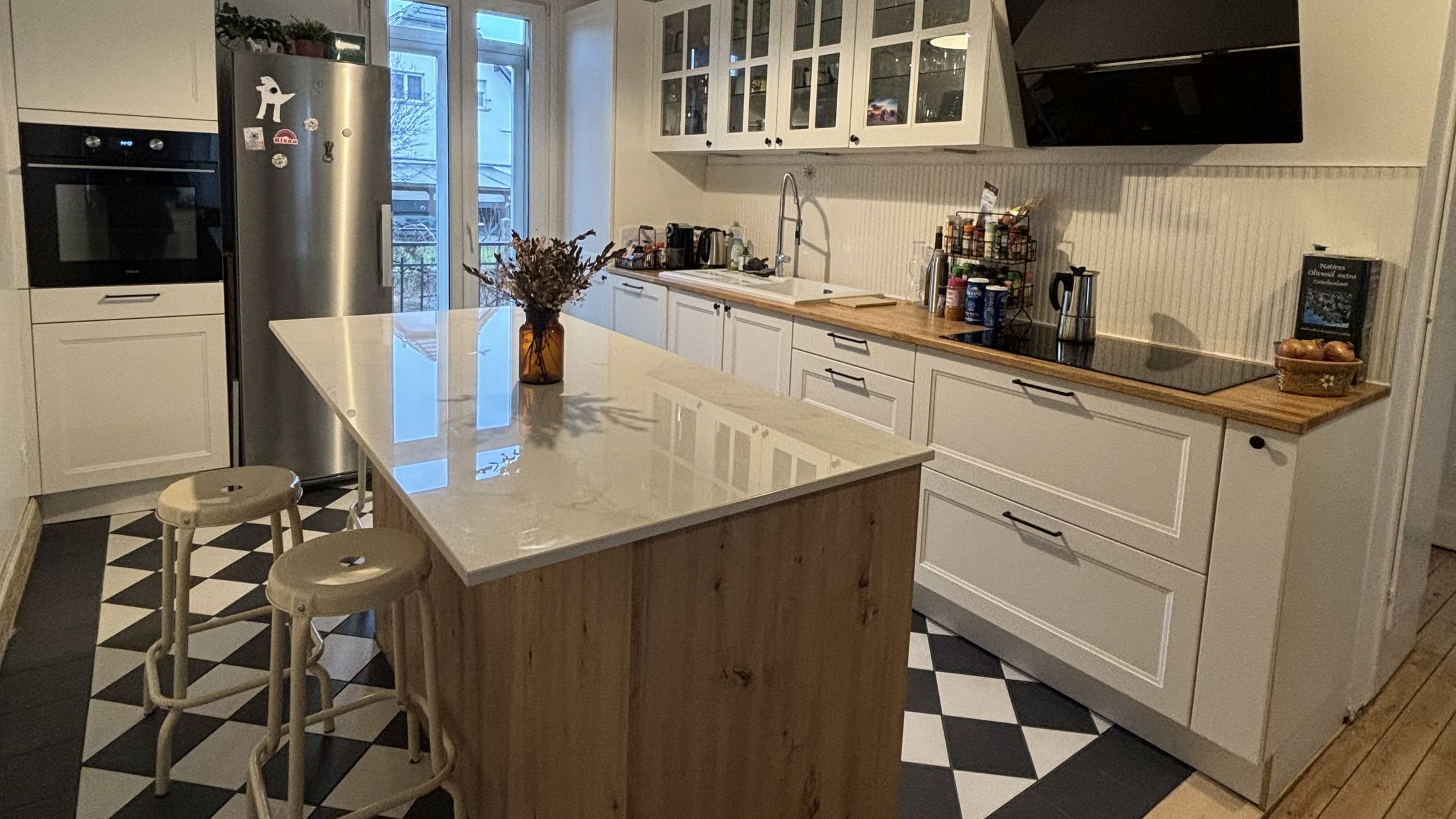 Vue d’ensemble : cuisine blanche à cadre avec meubles hauts vitrés et hotte inclinée noire, îlot central chêne à plan blanc, sol en damier noir et blanc bordé de parquet ancien
