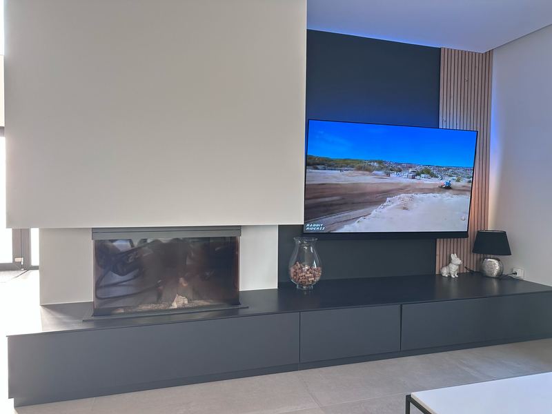 Mur TV : long meuble bas anthracite sans poignées intégrant un foyer de cheminée, téléviseur sur panneau sombre rétroéclairé, tasseaux bois verticaux