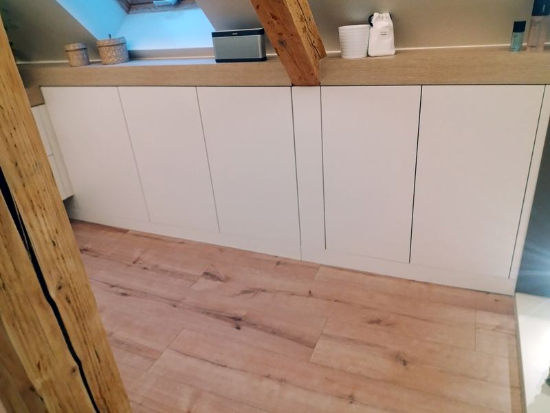 Rangements bas blancs sans poignées le long d'un rampant, tablette chêne, poutre de charpente apparente, sol décor chêne, fenêtre de toit