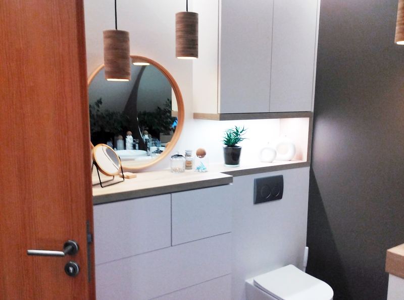 Salle d'eau vue depuis la porte : meubles bas blancs sans poignées avec plan décor bois, miroir rond cerclé de bois, deux suspensions cylindriques en bois, niche éclairée par LED sous meuble haut blanc, WC suspendu